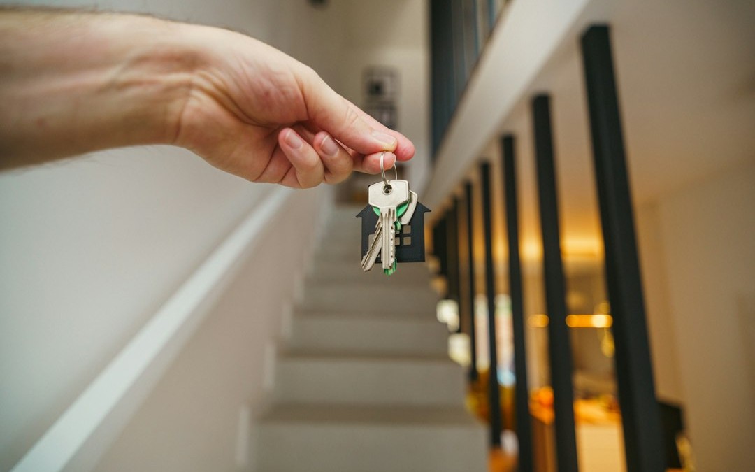 A hand holding keys with a house-shaped keychain in front of a staircase